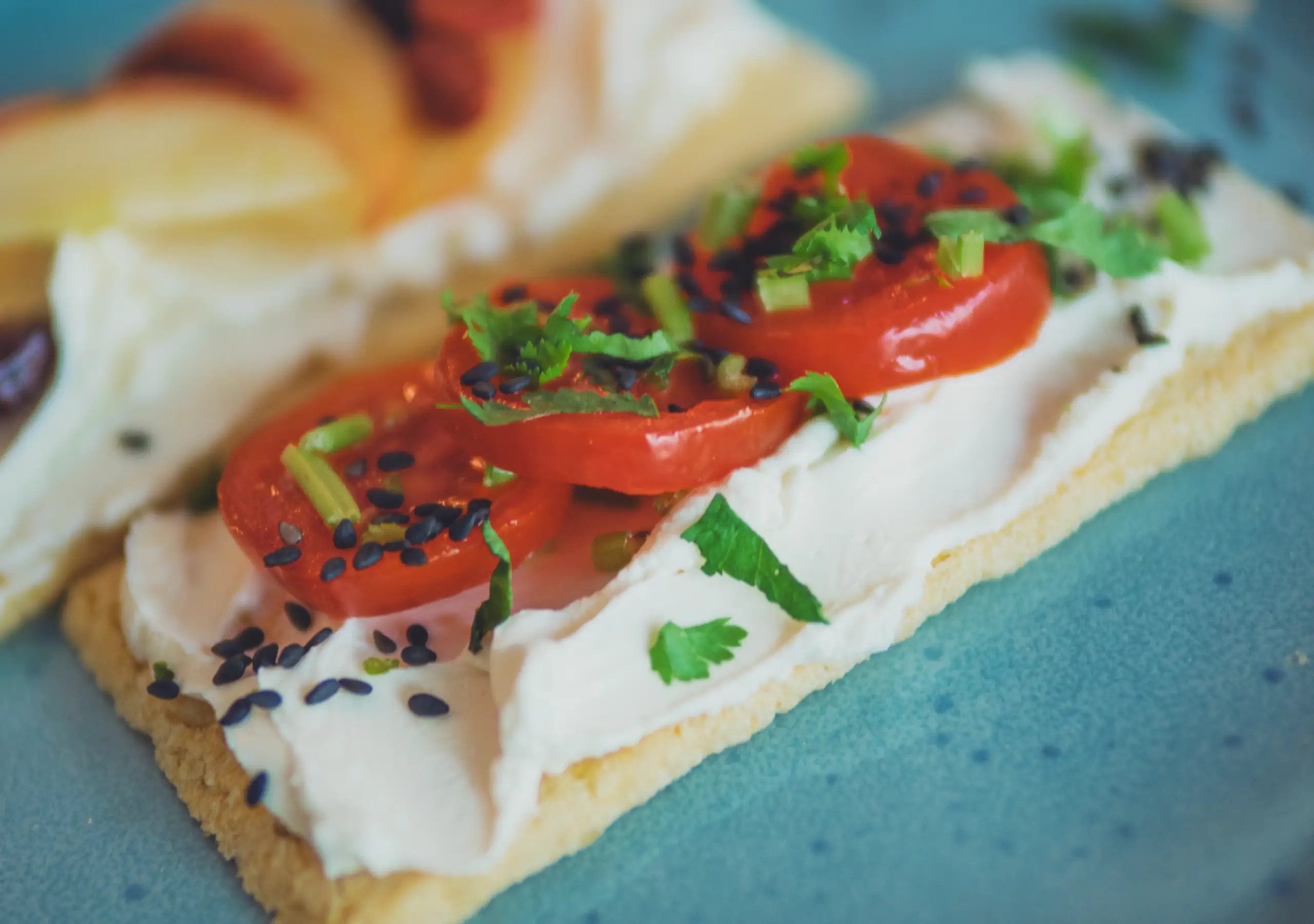 Nahaufnahme einer belegten Scheibe mit Frischkäse, Tomatenscheiben, Kräutern und schwarzem Sesam.