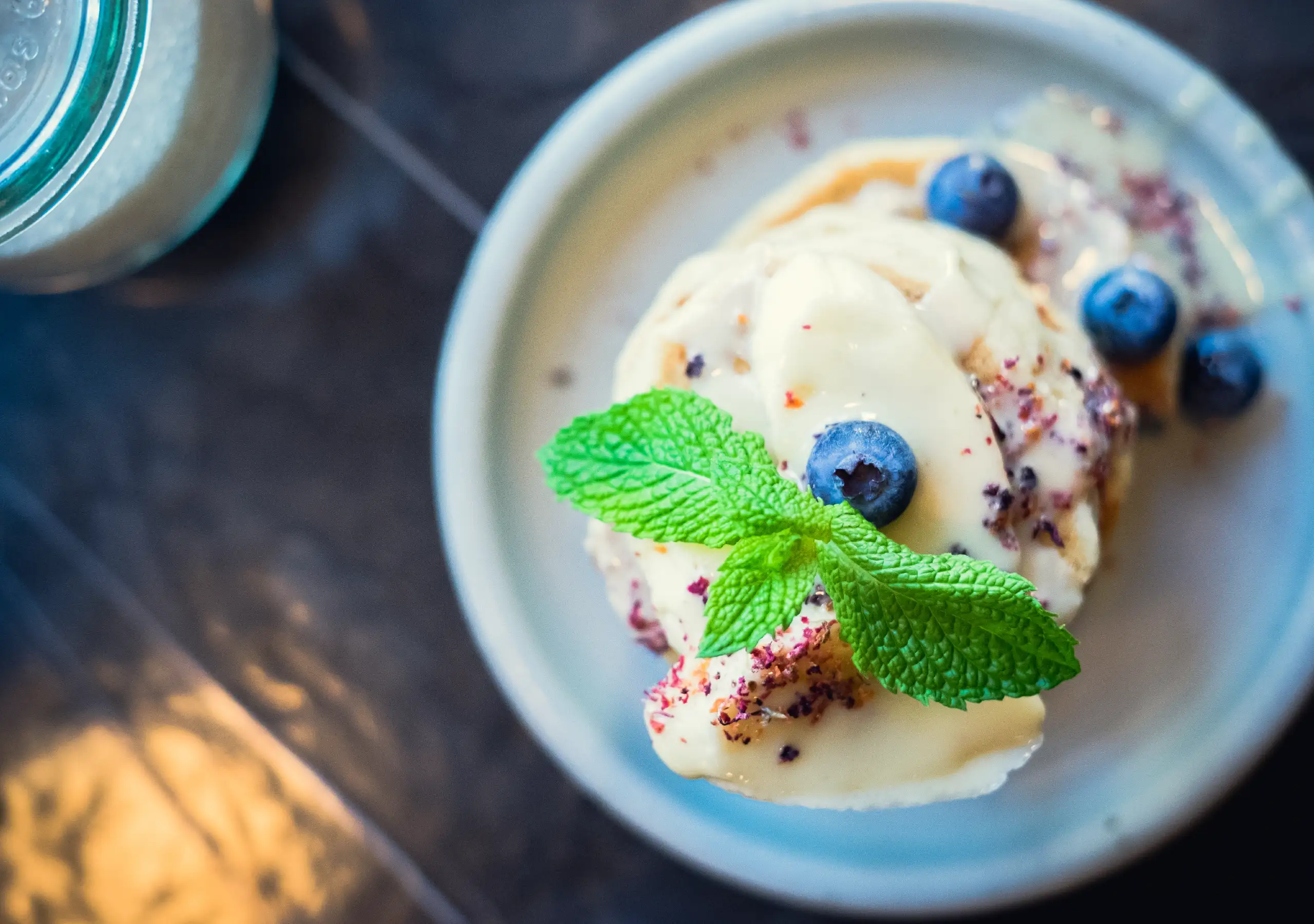 Teller mit fluffigen Pancakes, Blaubeeren, heller Sauce und frischer Minze in Nahaufnahme.