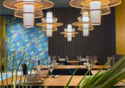 Mehrere runde, geflochtene Pendelleuchten vor dunklen Holzlamellen, gedeckte Tische.