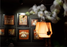 Wand mit gerahmten Illustrationen, warme Leuchten, unscharfe Blüte im Vordergrund.