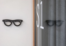 Piktogramm „Sehen“ – Raumkennzeichnung S1 6 Schwarzes Brillen-Piktogramm an Wand neben Glastür S1.