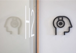 Piktogramm „Kopf/Diagnostik“ – Raum H2 4 Schwarzes Kopf-Icon mit Sensorik neben Glastür H2.